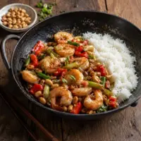 Kung Pao-style prawns with roasted peanuts, red pepper, spring onion and rice, served in a black wok on a rustic wooden table.
